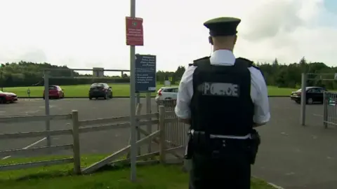 A police officer stands with his back to the camera at Henry Jones Playing Fields. He is wearing a hat, a dark bullet-proof/stab vest, dark trousers and a pale shirt. A number of cars are parked in the car park and there is a large grassed area in the background.