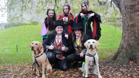 Guide Dogs Two Labradors with five Halloween characters dressed in torn black suits and colourful adornments 