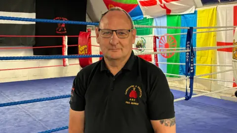 A picture of coach Paul Murray stood in front of a boxing ring. He has black glasses and is wearing a black polo shirt. He has tattoos on his arm and the boxing ring is blue in colour.