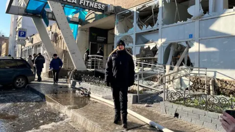 A woman wearing a blue jacket and hat stands in front of a damaged building