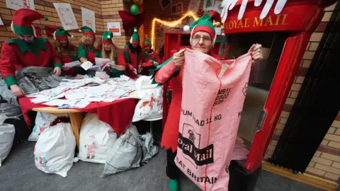 PA Media A woman dressed as a Christmas elf holds a large pink Royal Mail sack. In the background, a group of elves is gathered around a table sorting through letters. They all have green and red tunics on and green and red pointy hats adorned with large pointy elf ears. 