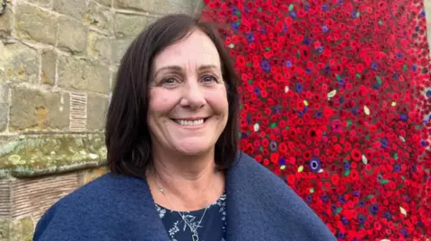 ELLEN KNIGHT/BBC Donna pictured looking into the camera and smiling. She's wearing a navy blue dress with a white floral pattern, and a lighter blue wool coat. She's stood in front of the bottom of the cascade - a wall of knitted poppies in red and purple. 