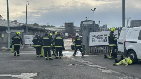 The image shows a eight uniformed members of the fire service standing outside the entrance to Plymouth Fisheries.