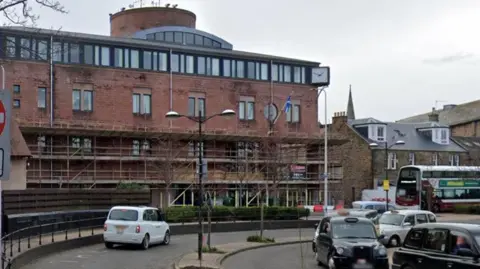 Midlothian Council offices - a red sandstone building with scaffolding along thr front. Cars drive on the road in front of the building.