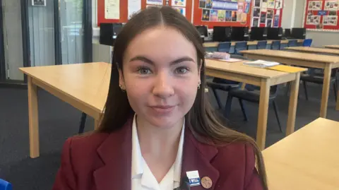 Ebony má dlouhé hnědé vlasy, které procházejí její ramena a ona se usmívá na kameru. Má na sobě kaštanovou školní bundu se dvěma kolíky na klopě a také bílou límečkovou košili. Sedí v prázdné třídě, s řadou dřevěných stolů za ní a červené desky jsou na zdi kolem učebny. Za ní je řada stolních počítačů.