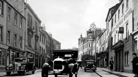Black and white photograph of old cars driving down a street made of stone buildings.