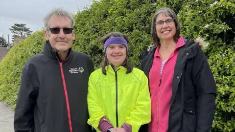Serena has brown hair and a purple sweatband on her head. She is wearing a bright yellow windbreaker and is smiling. She is standing in front of a green bush on a path. To her left is her dad who has grey hair and tinted black glasses, wearing a black jacket with a red zip. On the right is a woman with brown and grey hair. She has a pink top with a black coat on top