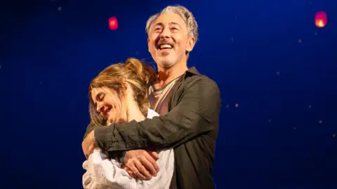 Two performers stand together on a dimly lit stage, framed by a deep blue backdrop and glowing red lanterns suspended overhead.
Warm spotlighting highlights the embrace, while the floating lights create a dreamlike, theatrical atmosphere.