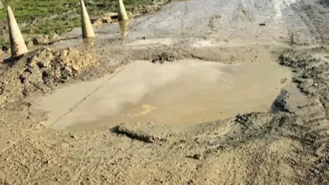 Sharon Oertel A large muddy hole can be seen on a road with muddy cones around it.