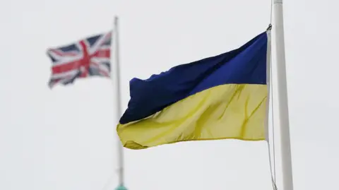 A blue and yellow Ukrainian flag flies, with a Union Jack in the background. It is a cloudy day.