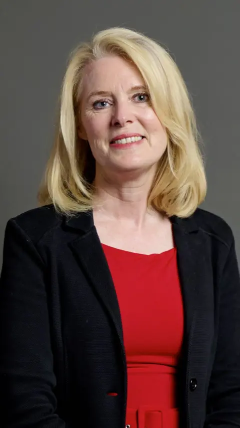Woman with blond hair smiles for camera. She wears a black jacket over a red dress