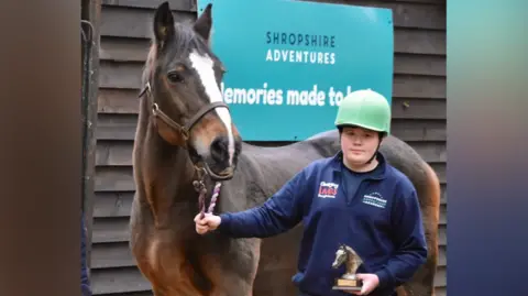Shropshire Adventure Academy A man wearing a blue jumper and trousers is stood infront of a brown and white horse holding a trophy in the shape of a horse's head. He is wearing a green helmet.