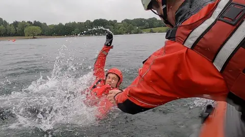 A firefighter wearing a red dry suit and floating in a lake is grabbed by another firefighter on a nearby boat