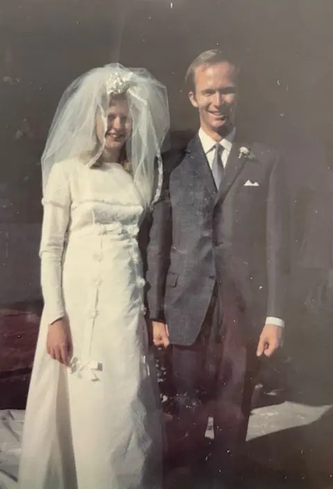 Foto de familia Barbie y Peter Reynolds tomados de la mano y sonriendo para una foto de retrato el día de su boda. Barbie está a la izquierda con un vestido de novia y un velo. Peter está de pie a su lado con un traje gris