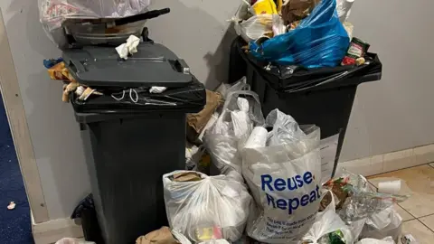 Handout Bins overflowing with rubbish and bags of rubbish lying on the floor in heaps with rubbish spilling out of them. 