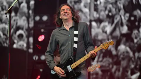 A man with long dark hair playing a black electric guitar.