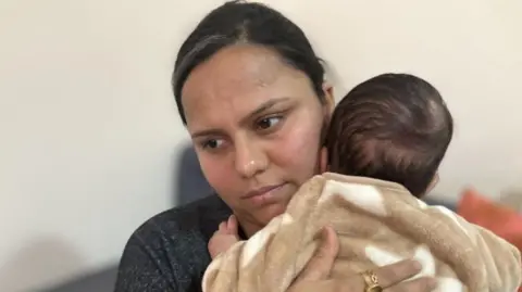 BBC Priya Rastogi is holding her baby in her arms. She has dark hair and is looking away from the camera.