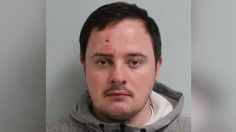 A young man wearing a grey hoody looks straight at the camera unhappily. He has short dark hair and a moustache. He also has two fresh scabs and dried blood  on his forehead above his right eye. 