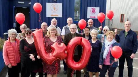 A group of men and women standing together in front of a blue metal-looking building while holding red balloons, A man and a woman in the middle of the group are holding up a "4" and a "0" balloon. They are all smiling and looking into the camera.