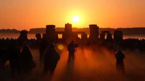 A hazy orange image showing the sun rising above the horizon at Stonehenge. A large crowd of people have gathered beneath the base of the stones, only their silhouettes are visible. A photographer stands next to a tripod to capture the moment. 