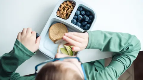 A school pupil pictured from above eating lunch wearing a green jumper and blue glasses. In the lunchbox in front of them there are some walnuts, blueberries, and some crackers with cream cheese and avocado. 