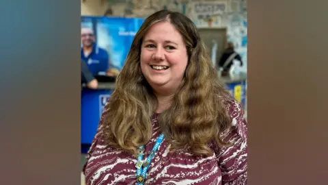Tanya Stubbs Tanha stubbs wearing a red top, she has long brown hair and is wearing an NHS lanyard. She is smiling towards the camera, and is pictured in a hospital setting