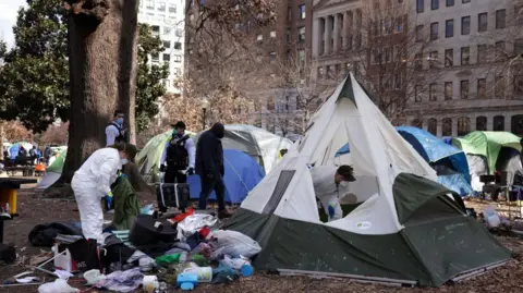 Getty Images A tent encampment being removed from downtown Washington DC in 2023. Workers in hazmat suits wear face masks. Rubbish is seen strewn outside of one white and green tent. There are large trees and buildings visible outside of the edges of the public park.