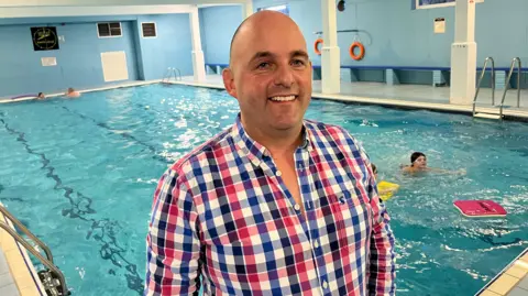 John Devine/BBC Julian Frost has a balding head and is wearing a red, blue and white gingham shirt. Behind him is a swimming pool with people in the water.
