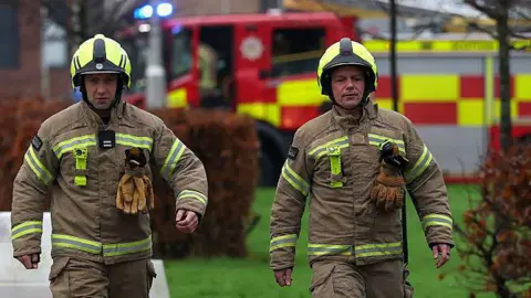 Getty Images Two male firefighters in full firefighting kit walk towards the camera, a fire appliance behind them in the background with its blue lights flashing.