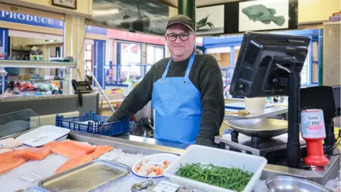 Kenny Brown Dave Walker, wearing a blue apron and green jumper, smiles behind the counter with salmon and other fish in front of him