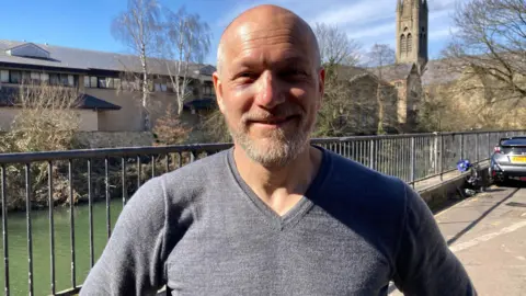 A head and shoulders picture of Dunstan Bertschienger. He is wearing a grey v-necked jumper and is smiling directly into the camera. He is standing by a river.