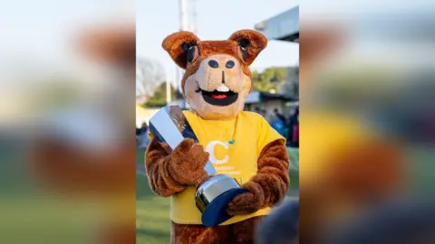 Lyn & Trev Sports A person in an animal suit, wearing a yellow t-shirt and holding a silver trophy. 