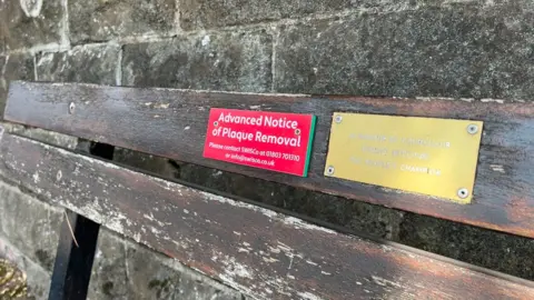 A plaque in red and white warns of the removal of the golden plaque next to it honouring former councillor Fred Stone