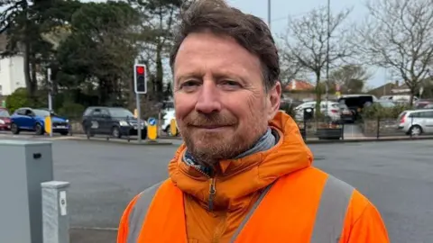 Jersey Highways manager Tristen Dodd is wearing a high vis orange jacket with silver reflective stripes on it. He has a lighter orange coat on underneath and is looking at the camera. He has brown hair which is shorter on the sides and longer on top and is standing beside a four-way traffic light controlled junction with some vehicles visible in the background.