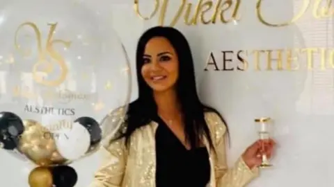 A person stands in a bright, modern interior beside a white wall featuring a gold logo and the text “AESTHETICS.” The person is wearing a black outfit with a gold belt and a gold sequined jacket, holding a large transparent balloon