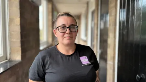 Martin Giles/BBC Shauni Stannard smiles in a corridor. She has dark hair that has been tied into a bun and she wears a black T-shirt. 