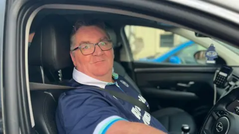 A man in a short-sleeved Scotland rugby top with dark hair and glasses sits at the wheel of a car with his seat belt on