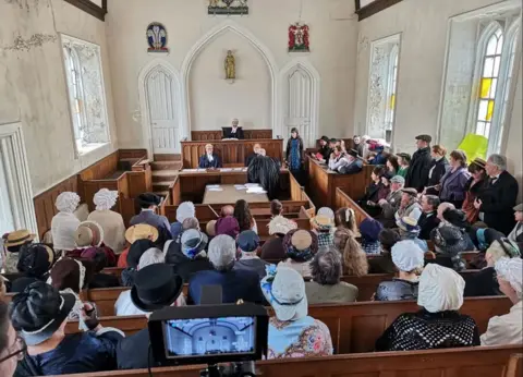 A camera in the foreground films a scene from The Ballad of Lucy Sands. A large number of people wearing Victorian clothes sit in a room set up as a court, with judges seated at the far end. The building resembles a chapel, with large stained-glass windows on either side.