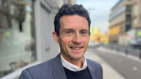 A man with short dark hair smiling while standing on a high street. He is wearing a grey suit jacket, navy jumper and white shirt.