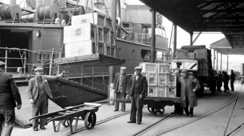 A black and white photo of Bristol docks with a large container ship unloading large crates onto trolleys, with several men in flat caps in the foreground