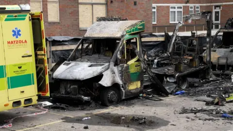 An ambulance damaged by fire in a car park. More than half of the vehicle has come apart and there is soot all over. The floor nearby is black and there is clutter everywhere. There is another damaged vehicle on the right of it covered in soot. The ambulance on the left-hand of the image looks untouched by the fire and has Hatzola Northwest on the back door.