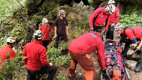 SARA Eight members of Severn Area Search and Rescue wearing red uniforms and white helmets. They are pictured during a training exercise, tending to a woman in a stretcher in the middle of some woodland in the Lower Wye Valley near Tintern.