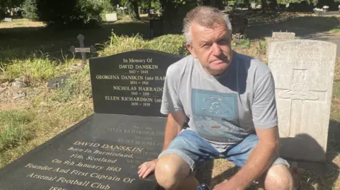 Ian Woolley, a man in jean shorts and a grey graphic tshirt kneels next to a black gravestone which reads David Danskin 1863-1948
