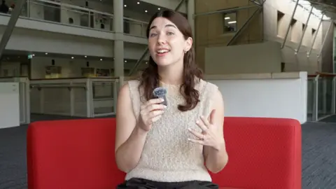 BBC journalist Claire Keenan sitting on a red couch holding a small microphone