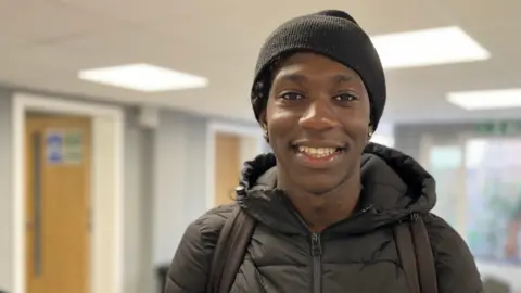 Jon Wright/BBC A young black boy, with a coat and hat, standing in large room smiling at the camera