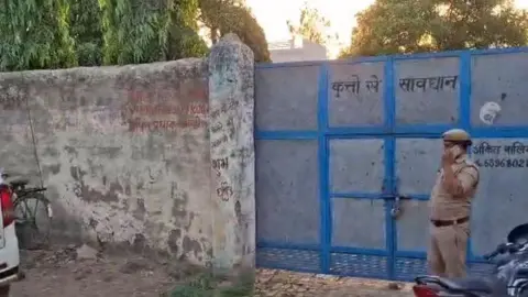 BBC Hindi A policeman standing outside a grey metal gate which says beware of dogs 