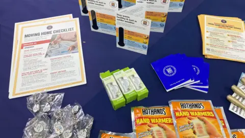 A table covered with a blue tablecloth, displaying individual piles of hand warmers, hour glasses for reducing time spent in the shower, keychains wrapped in plastic, and information booklets.