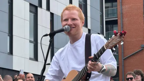 Zoie O'Brien/BBC Ed Sheeran performing with his guitar at Ipswich Waterfront last week. He is standing singing in front of a microphone and mic stand and is wearing a white T-shirt.