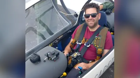 Richard Smith, a man who is looking directly at the camera and smiling. He is sitting in a plane and is wearing a red T-shirt and a pair of sunglasses.