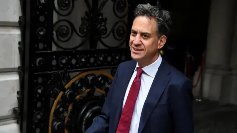 Reuters Ed Miliband walking out of a large ornately-decorated iron gate outside Downing Street in London. He is wearing a dark blue suit, white shirt and red tie.